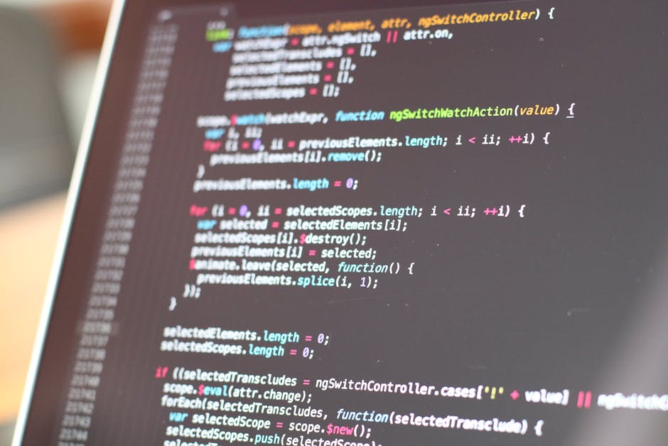 A close-up view of a person's hand typing on a laptop keyboard placed on a wooden surface. The laptop screen displays lines of code with syntax highlighting in various colors, indicating programming a