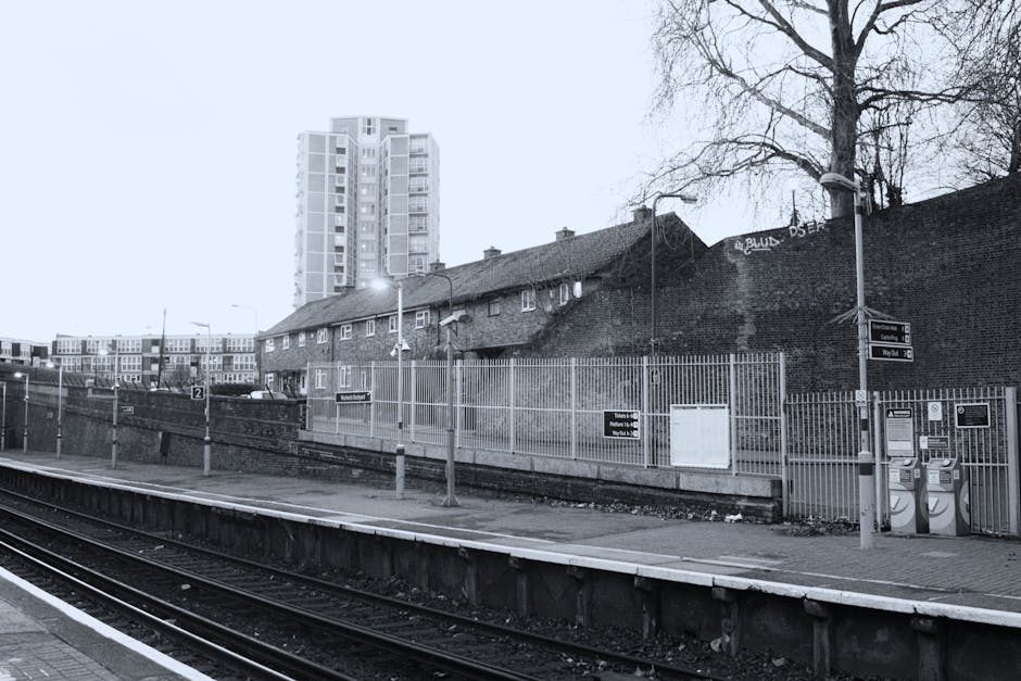A black and white photograph of a train station platform in an urban setting. In the foreground, there are railway tracks with wooden sleepers and metal rails. The platform is edged with a concrete cu