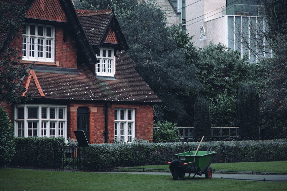 A traditional red-brick house with a steep, tiled roof and white-framed sash windows situated in a well-maintained garden. The garden features a neatly trimmed hedge border, lush green grass, and matu